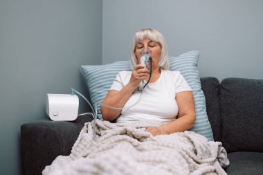 Kafkasyalı yaşlı bir kadın astım spreyiyle hastalandı. Sağlıksız bir kadın evde solunum yapıyor. Tedavi için nebulizör ve sprey kullanıyor. Evdeki kanepede oturuyor.. 