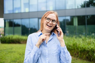 Neşeli, mutlu, şık bir kadın alışveriş merkezinin yanında dururken akıllı telefon uygulaması kullanıyor. Mavi tişörtlü mutlu kadın caddede yürürken akıllı telefonuna bakıyor.