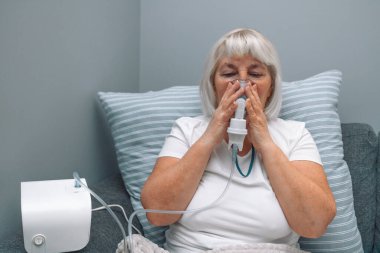 Kafkasyalı yaşlı bir kadın astım spreyiyle hastalandı. Sağlıksız bir kadın evde solunum yapıyor. Tedavi için nebulizör ve sprey kullanıyor. Evdeki kanepede oturuyor.. 