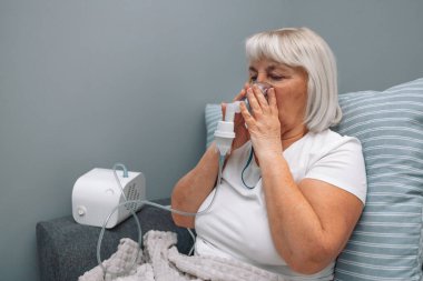 Kafkasyalı yaşlı bir kadın astım spreyiyle hastalandı. Sağlıksız bir kadın evde solunum yapıyor. Tedavi için nebulizör ve sprey kullanıyor. Evdeki kanepede oturuyor.. 