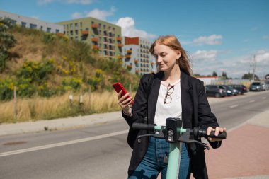 Akıllı toplu taşıma aracı. Mutlu 30 'lu yaşlardaki genç bir kadın elektrikli scooter kiralıyor, akıllı telefon uygulaması ile çevre dostu ulaşımı açıyor, açık bir şehir bölgesindeki bisiklet parkının yanında duruyor.