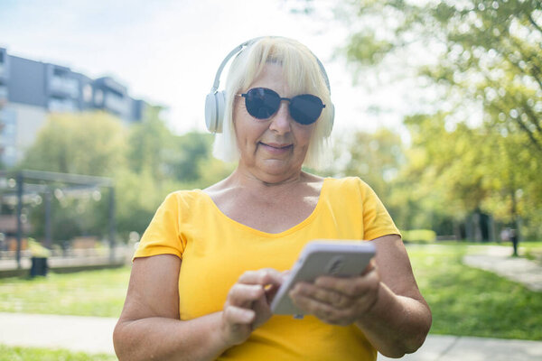 Cheerful caucasian mature woman wife using smart phone cellphone, choosing sound track online while listening to the music in headphones in park. High quality photo