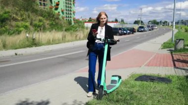 Akıllı toplu taşıma aracı. Mutlu 30 'lu yaşlardaki genç bir kadın elektrikli scooter kiralıyor, akıllı telefon uygulaması ile çevre dostu ulaşımı açıyor, açık bir şehir bölgesindeki bisiklet parkının yanında duruyor.