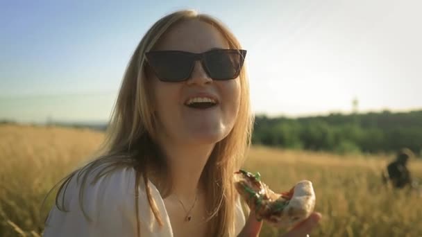 若い美しい陽気な少女は 夕日ピクニックで自然の味を楽しんでピザを食べています ピザと友人は ライフスタイルの食べ物のための街並みで屋外を食べる — ストック動画