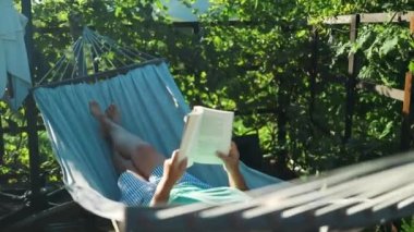 Güzel sarışın kadın kitap okuyor ve bahçedeki hamakta dinleniyor. Yüksek kalite 4k görüntü