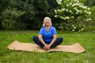 Atletik 50 'lerin son sınıf öğrencisi kadın yoga minderinde poz veriyor. Spor kıyafetleri giymiş, parkta ya da ormanda tek başına. Yüksek kalite fotoğraf