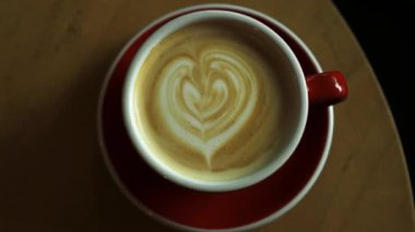 Sıcak kahvenin üst görüntüsü ve ahşap masa arkasındaki kırmızı bardakta kalp şeklinde latte sanatı. Yüksek kaliteli FullHD görüntüler