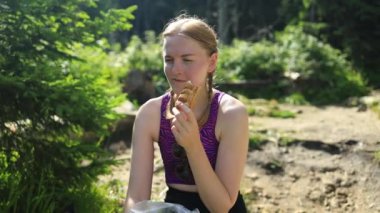 Bir kayanın üzerinde oturan yorgun, beyaz bir kadın yürüyüş sonrası doğada sandviç yemeye hazırlanıyor. Yüksek kaliteli FullHD görüntüler