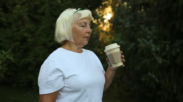 外でコーヒーや紅茶の紙コップを持っている幸せな50代の金髪の女性の肖像画 彼女は夏に彼女自身で夜を楽しんでいる 高品質のフルHd映像 — ストック動画