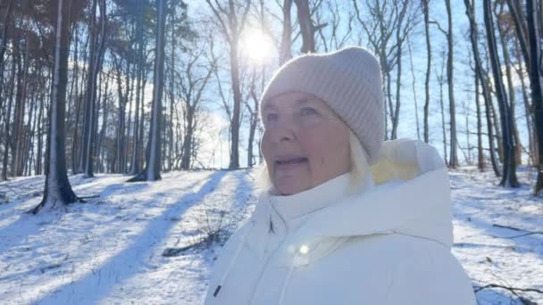 木や雪の漂流の背景に屋外のカメラを見ながら暖かい帽子で笑顔で魅力的な50代の上級女性のクローズアップ肖像画 雪だるま市立公園でポーズする幸せな女性のクローズアップヘッドショット — ストック動画