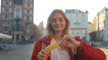 Avrupa sokaklarında muz meyvesi yiyen 30 'lu yaşlardaki beyaz bir kadının açık hava spor portresi. Krakow, Polonya. Yüksek kaliteli FullHD görüntüler
