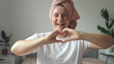 Romantik, mutlu, beyaz kadın portresi. Evde kameraya gülümserken elleriyle kalp şekillendiriyor. Yüksek kaliteli FullHD görüntüler