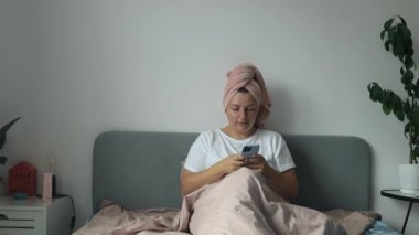 Genç bir kadın yatakta cep telefonu kredi kartı alışverişi yaparak pijama giyer rahatça yatak odasında vakit geçirir. Yüksek kaliteli FullHD görüntüler
