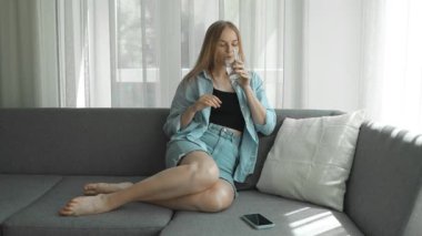 Bir bardak su tutan kız. Oturma odasındaki kanepede su içen gülümseyen kız. Yüksek kaliteli FullHD görüntüler