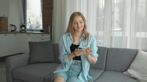 快乐的30多岁的金发女孩拿着手机在家里用智能手机装置 笑笑年轻的白人女性博主订阅新的社交媒体 在网上购买 在线订购应用程序产品 — 图库视频影像