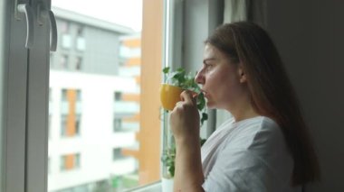 Bir fincan çay ya da kahveyle rahatlamış güzel bir kadın. Pencere kenarında otururken pencereden bakıyor. Yüksek kaliteli FullHD görüntüler