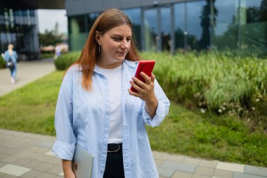 Neşeli, mutlu, şık bir kadın alışveriş merkezinin yanında dururken akıllı telefon uygulaması kullanıyor. Mavi tişörtlü mutlu kadın caddede yürürken akıllı telefonuna bakıyor.