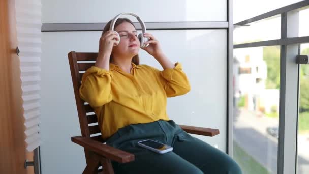 若い美しい白人女性は テラスに座っている間 ヘッドフォンを使用してリラックスして音楽を聞きます — ストック動画