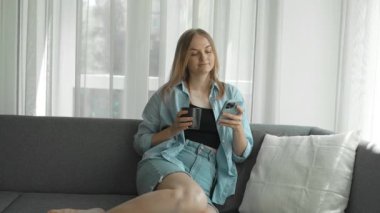 30 'lu yaşlardaki rahat bir kadın akıllı telefon kullanıyor ve kanepede otururken fincandan kahve içiyor. Milenyum kızı evinde cep telefonu teknolojisiyle vakit geçiriyor. Yüksek kaliteli FullHD görüntüler