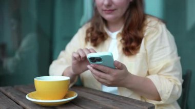 30 'lu yaşlarda mutlu, beyaz bir kadın sarı gömlek giyiyor cep telefonuyla sohbet ediyor çay içiyor kahve dükkanındaki masada tek başına oturuyor. 