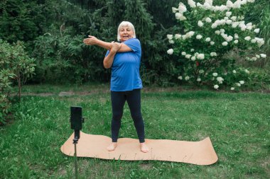 50 'li yaşlardaki kadın yoga minderinde pratik yapar, akıllı telefon kamerasında konuşur, takipçiler için video içeriği oluşturur, dışarıdaki tripodlarda cep telefonundaki webcam' e bakar. Spor Blog yazarı kavramı. 