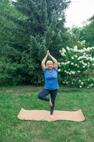 Yoga at park.Full length shot of happy energetic mature 50s woman in casual clothes exercising outdoors, practicing yoga on mat, standing in warrior ii pose. Age, wellness and health. Healthy