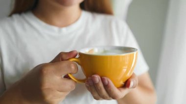 Bir kupa içecekle kadın elinin yakınlaşması. Güzel bir kız elinde sarı kapuçino fincanı ile sabah güneşinde kahve tutuyor.. 