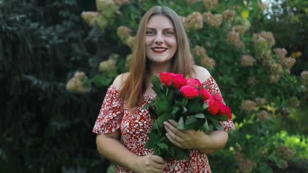 バラを保持する赤いドレスの美しい魅力的なスタイリッシュな女性の肖像画 屋外にポーズする花ロマンチックな気分 — ストック動画