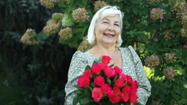 美しい魅力的なスタイリッシュな50代シニア女性の肖像画 バラを保持する赤いドレス 屋外にポーズする花ロマンチックな気分 — ストック動画
