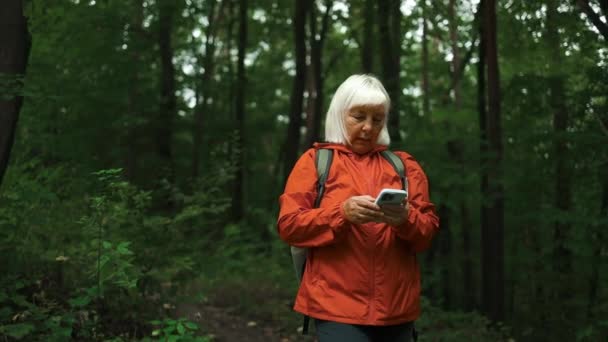 森で週末を楽しんでいる間 スマートフォンを使用してシニア女性ハイカー 携帯電話で森林での魅力的な老婦人バックパッカー計画旅行ルートの低い角度ビュー 高品質のフルHd映像 — ストック動画