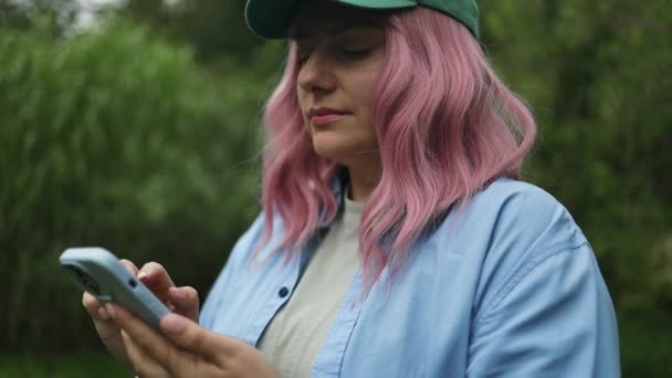 白人の若いかわいいピンクの髪の女性 通りを歩く緑の帽子の帽子の配達労働者 屋外で使用された携帯電話の高品質のフルHd映像 — ストック動画