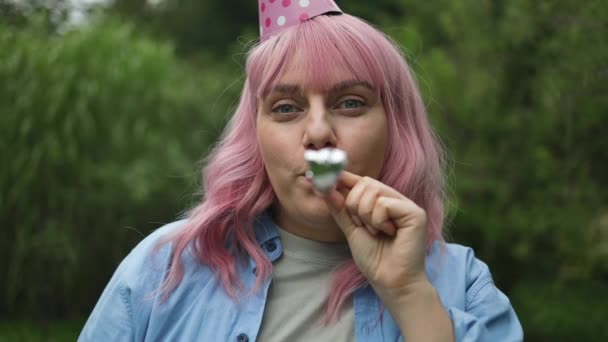 快乐有趣惊奇的年轻的粉红头发白种人女人头戴生日礼帽 手举着烟斗 看着后院的相机 — 图库视频影像