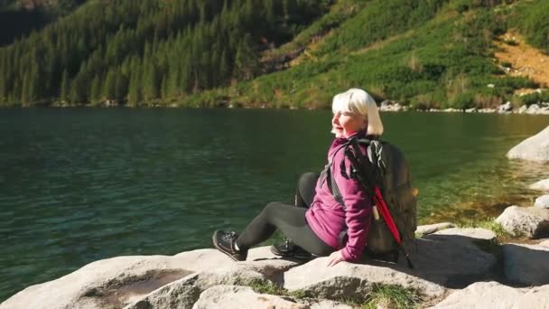 ポーランドのタトラ国立公園の山岳湖の背景に対して楽しんで岩の上に楽しむバックパックトレッキングとスポーツ服のシニア女性 Goal 自由と達成 — ストック動画
