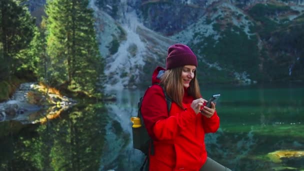 観光ハイカーの女性バックパッカーは ポーランドのタトラ国立公園のモラン オコ山湖の景色の近くでスマートフォンでセルフィーを取ることを楽しんで岩の上に アドベンチャー 休日のコンセプト — ストック動画