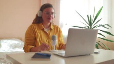 Dizüstü bilgisayarında kulaklıklar ve mikroskopla podcast yapan bir kadın. Kadın podcaster kendi stüdyosundan ses yayını yapıyor. Yüksek kaliteli FullHD görüntüler