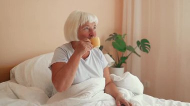 60 'lı yaşlardaki beyaz elbiseli neşeli bir kadın yatakta bir fincan kahveyle oturup kahvaltısını yaparken yatak odasının keyfini çıkarıyor. Yüksek kaliteli FullHD görüntüler