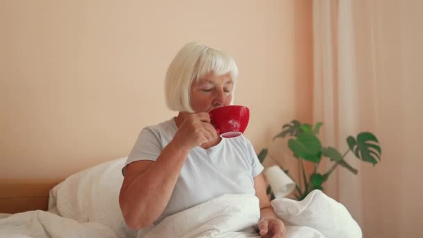 穿着白色睡衣的快乐的60多岁的女性坐在床上喝咖啡 一边吃早饭 一边在家里的卧室里享受着早晨的时光 优质Fullhd影片 — 图库视频影像