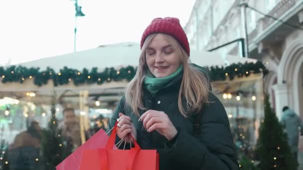 魅力的な幸せな白人ブロンドの女性は チャトリスタマーケットで赤い現在のショッピングバッグで通りを歩いています 雪の休日のクラクフの街で美しい女性の肖像画 高品質のフルHd — ストック動画