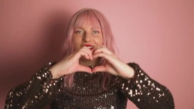 Elbise giymiş güzel beyaz kadın portresi kalp şeklinde el hareketleriyle pembe arka planda poz veriyor. Sevgililer Günü. Yüksek kaliteli FullHD görüntüler