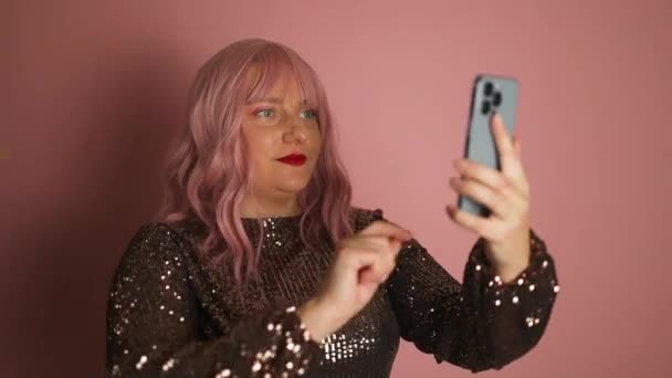 バレンタインデー 白い幸せなピンクの髪の女性は携帯電話を使用して光沢のあるドレスを身に着け ピンクの背景に隔離された笑いながらブログのためのビデオを記録します — ストック動画