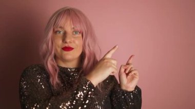 Genç, mutlu, neşeli, neşeli, başarılı Avrupalı, zarif bir elbise giyen, bir şeyi gösteren, gülümseyen ve pembe arka planda kendine güvenen bir kadın. 