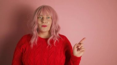 Genç, mutlu, neşeli, neşeli, başarılı, kırmızı kazaklı, bir şeye işaret eden, gülümseyen ve pembe arka planda kendinden emin görünen Avrupalı kadın. 