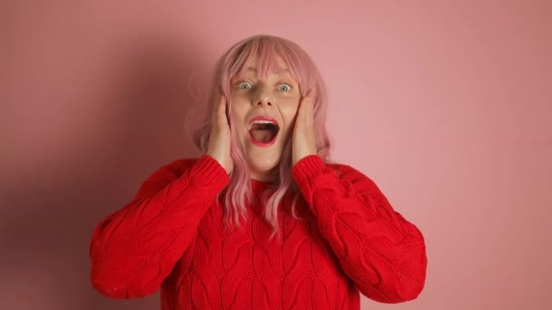 ヤングは カメラを見ながらピンクの背景に隔離された赤いセーターでピンクの髪の女性を驚かせた 高品質のフルHd映像 — ストック動画