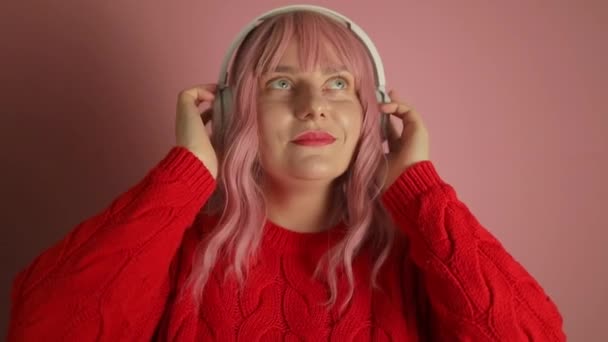 年轻快乐有趣的高加索女人穿着针织红毛衣耳机听音乐 在朴素的彩色浅粉背景图片上被隔离 人的生活方式概念 高质量的 — 图库视频影像