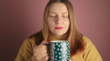 Mutlu bir kadın elinde bir fincan sıcak içecekle sabahları kahve ya da çay içerken pembe arka planda kapalı gözlerle eğleniyor. Yüksek kaliteli FullHD görüntüler
