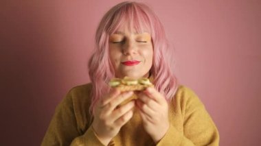 Mutlu, aç, pembe saçlı bir kadının stüdyo portresi. Muzlu lezzetli bir sandviç yiyor ve pembe izole edilmiş arka planda kameraya bakıyor.. 