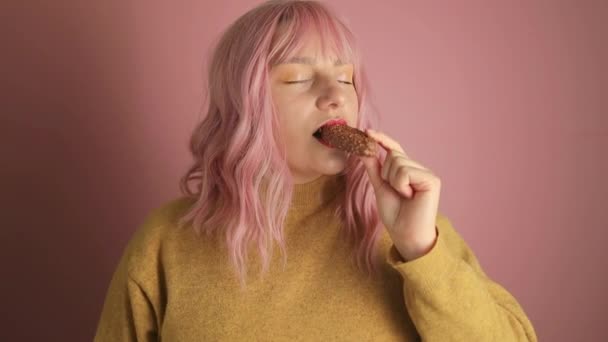 Счастливая Молодая Розововолосая Женщина Кусает Шоколадный Батончик Орехами Розовом Фоне — стоковое видео