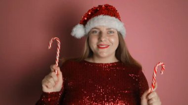 Noel Baba şapkalı, kırmızı elbiseli güzel bir kadın pembe arka planda baston şekerlerle kalp yapıyor. Noel kutlaması. Yüksek kaliteli FullHD görüntüler