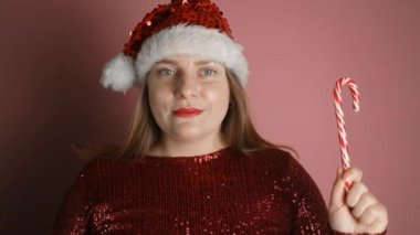 Noel Baba şapkalı, kırmızı elbiseli güzel bir kadın pembe arka planda baston şekerlerle kalp yapıyor. Noel kutlaması. Yüksek kaliteli FullHD görüntüler