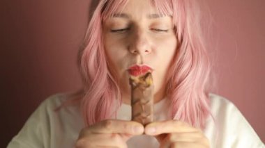 Gülümseyen, beyaz kazaklı, çikolata ısıran pembe saçlı genç kadın. Sade pastel açık pembe arka planda izole edilmiş bir kamera gibi görünüyor. İnsanların yaşam tarzı gıda konsepti. Yüksek kaliteli FullHD görüntüler